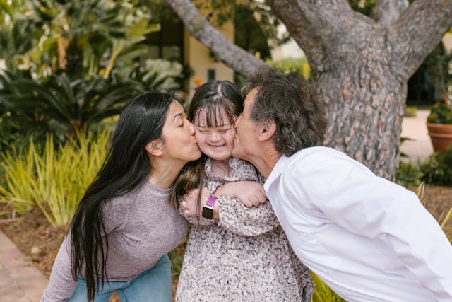 parents kissing their daughter with downs syndrome by a tree